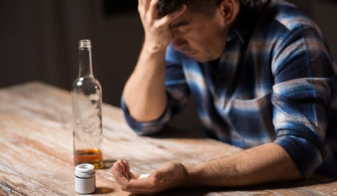 Мужчина с таблетками в руке и бутылкой алкогольного суррогата на столе
